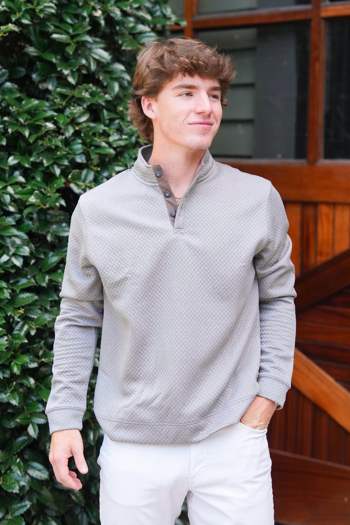 A young man wearing a light gray textured quarter-zip pullover sweater and white pants, standing outdoors in front of a green hedge and wooden staircase.