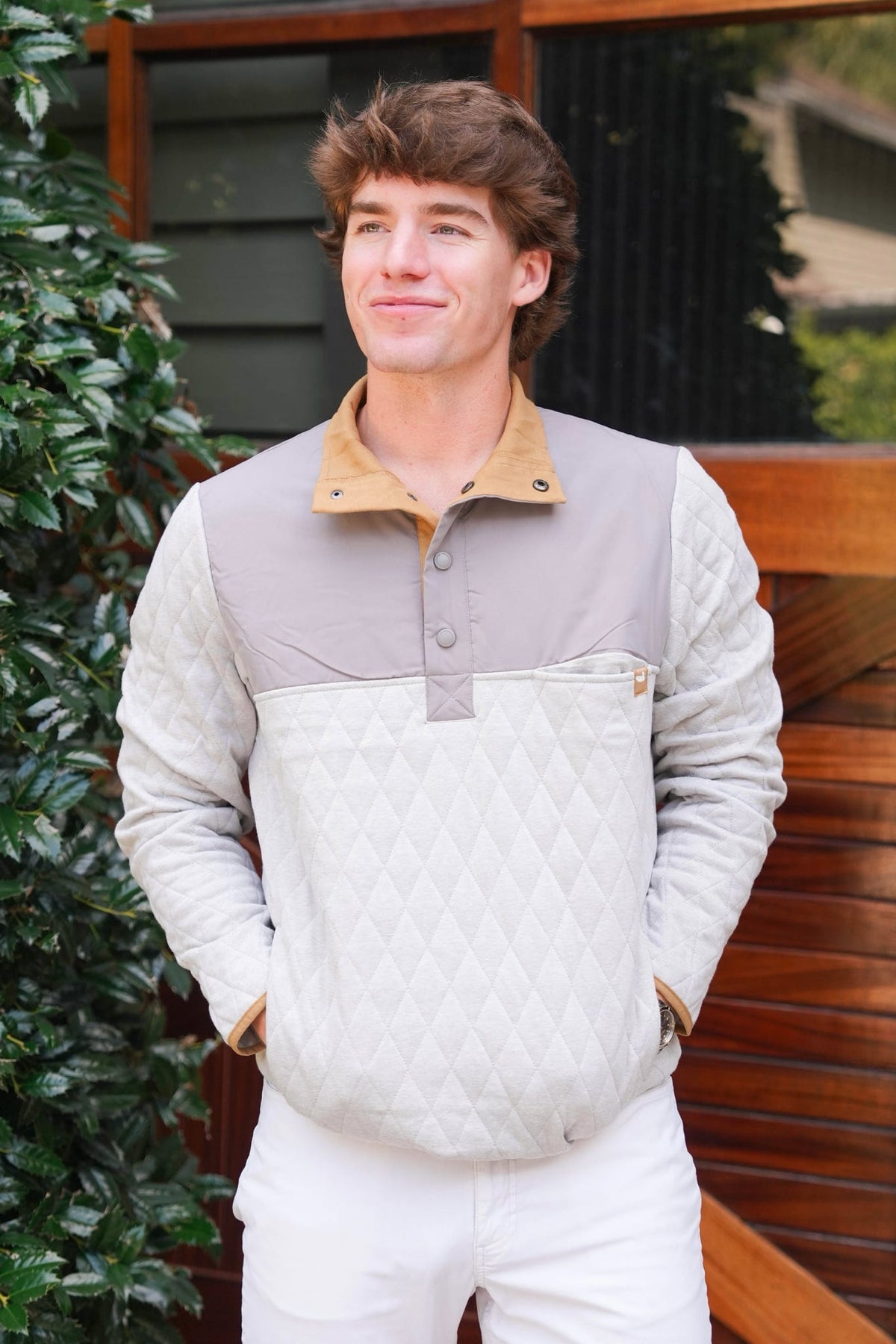 A man wearing a color-blocked quilted pullover jacket with a tan collar and white pants, standing outdoors in front of a wooden deck and greenery.