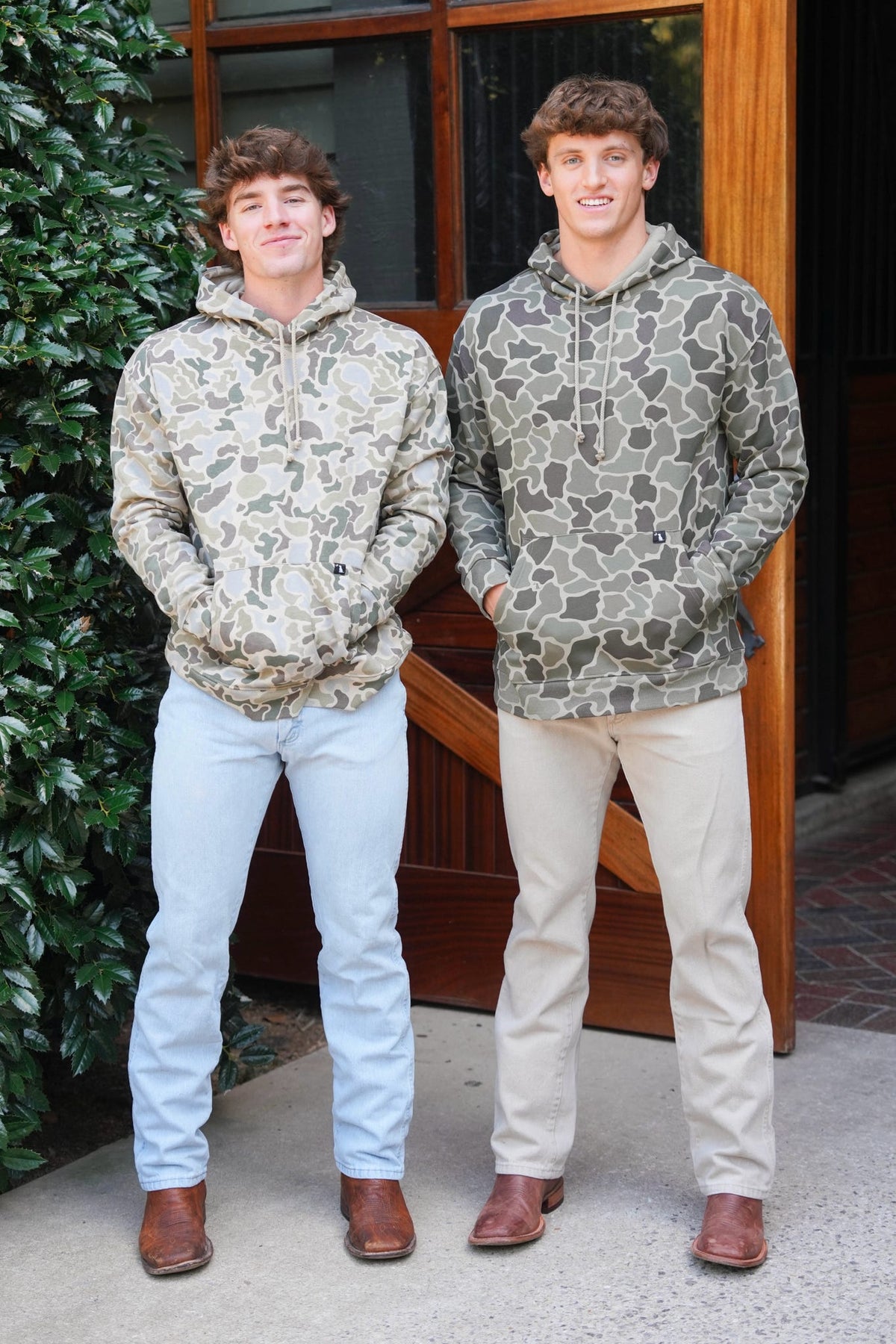 Two young men standing side-by-side wearing matching camouflage hoodies, with one in light blue jeans and the other in beige jeans, standing in front of a wooden door.