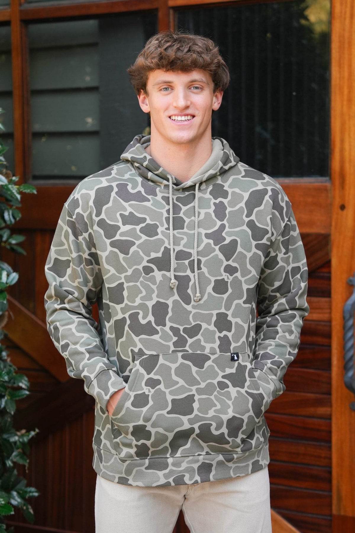 A smiling young man wearing a camouflage hoodie with a gray and olive green pattern, standing in front of a wooden door.