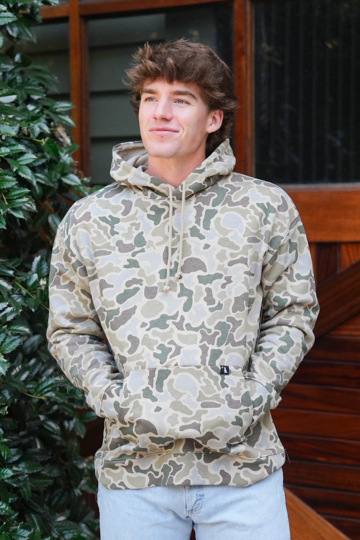 A man wearing a camouflage pullover hoodie with a kangaroo pocket, standing outdoors in front of a wooden door and green foliage.