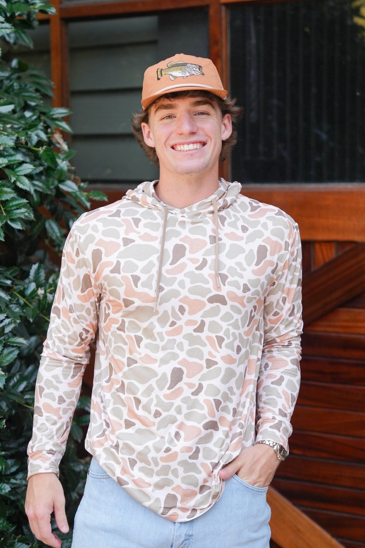 A smiling man wearing a beige and brown abstract patterned long-sleeve hoodie, a tan baseball cap, and light blue jeans, standing outdoors in front of a wooden door and green foliage.
