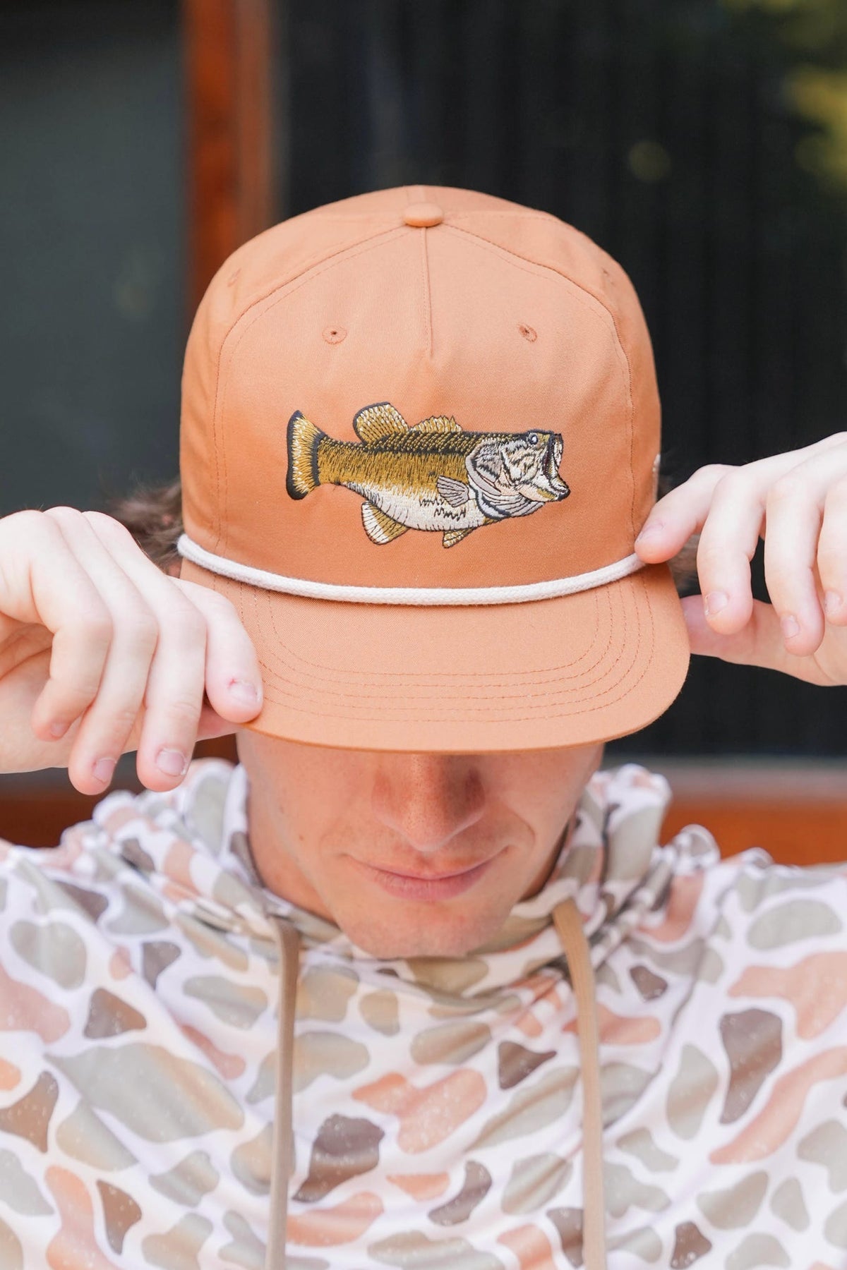 A person wearing a peach-colored baseball cap with an embroidered bass fish on the front, holding the brim down with both hands. The person is also wearing a patterned hoodie.
