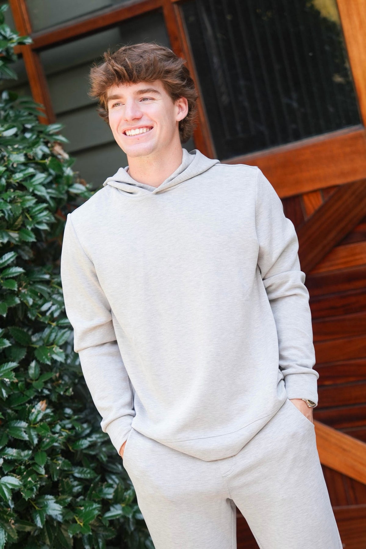 A smiling young man wearing a light gray heathered hoodie and matching sweatpants, standing outdoors in front of a wooden door and green foliage.