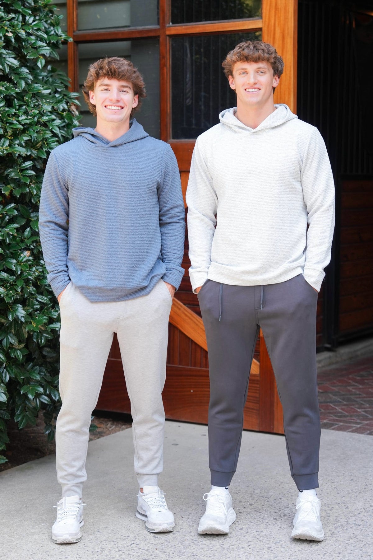 Two young men standing outdoors in front of a wooden door, wearing casual hoodies and sweatpants. The man on the left wears a slate blue hoodie with light beige joggers, and the man on the right wears a light gray hoodie with dark gray joggers.