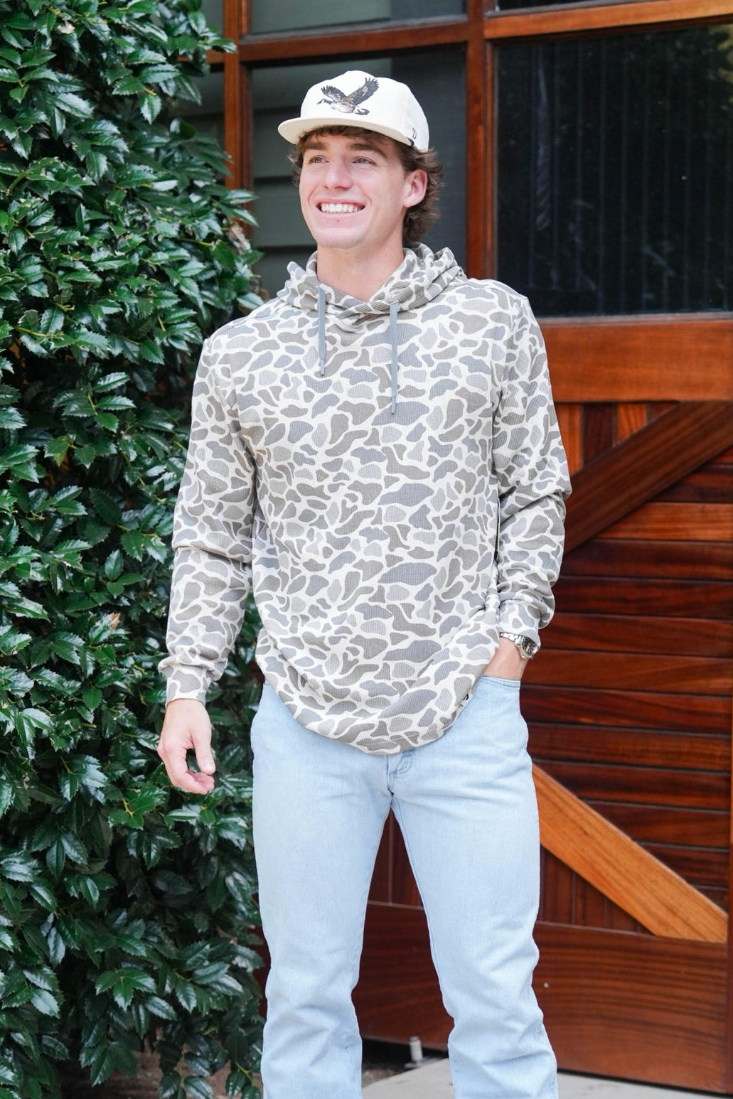 A man wearing a beige and brown animal print hoodie, light blue jeans, and brown boots stands in front of a rustic wooden door and green bushes.