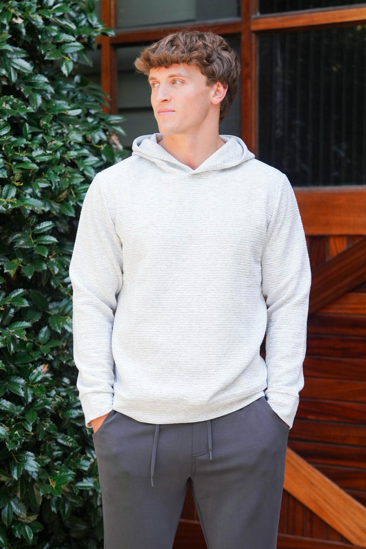 A man wearing a light gray textured hooded sweatshirt and dark gray sweatpants, standing outdoors in front of a wooden door and green foliage.