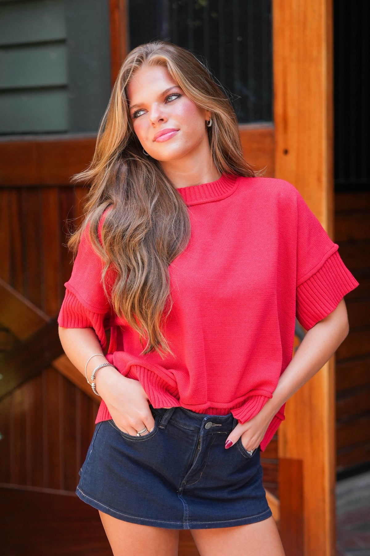 A woman wearing a bright coral-red short-sleeve knit top and a dark denim mini skirt, standing in front of a wooden door.