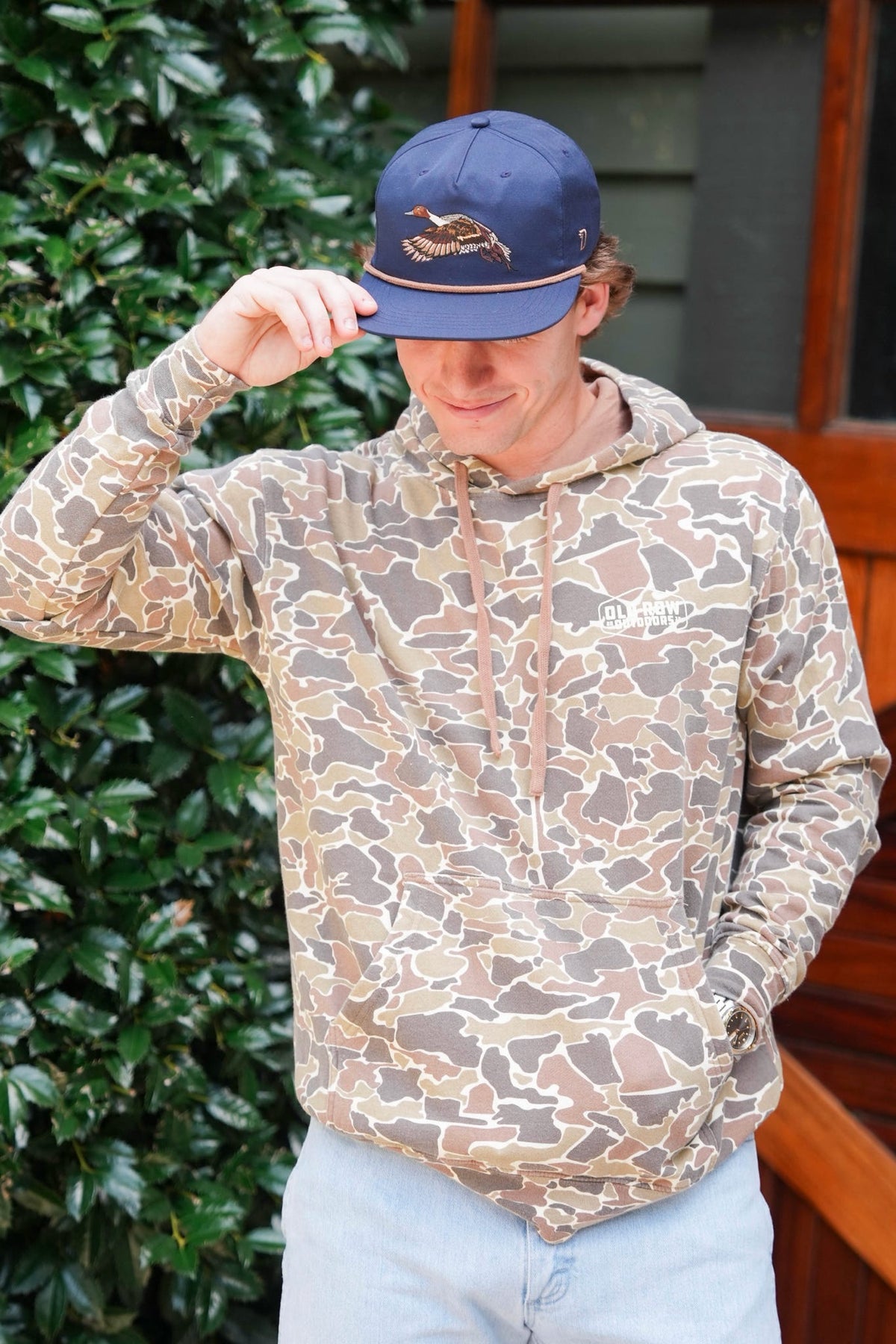 Person standing outdoors in a tan camouflage hoodie and navy cap embroidered with a flying duck, smiling while holding the cap’s brim.