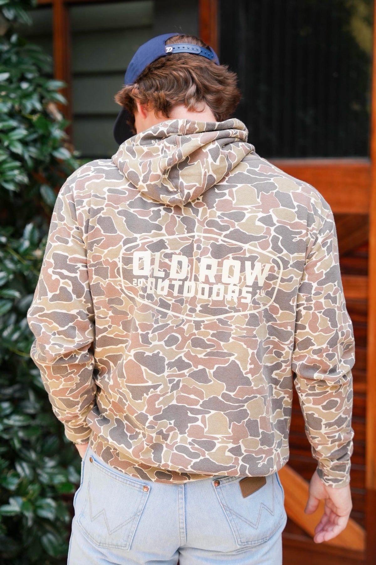 A man viewed from behind wearing a brown and beige camouflage hoodie with 'OLD ROW OUTDOORS' printed in white across the back, paired with light blue jeans and a navy blue cap.