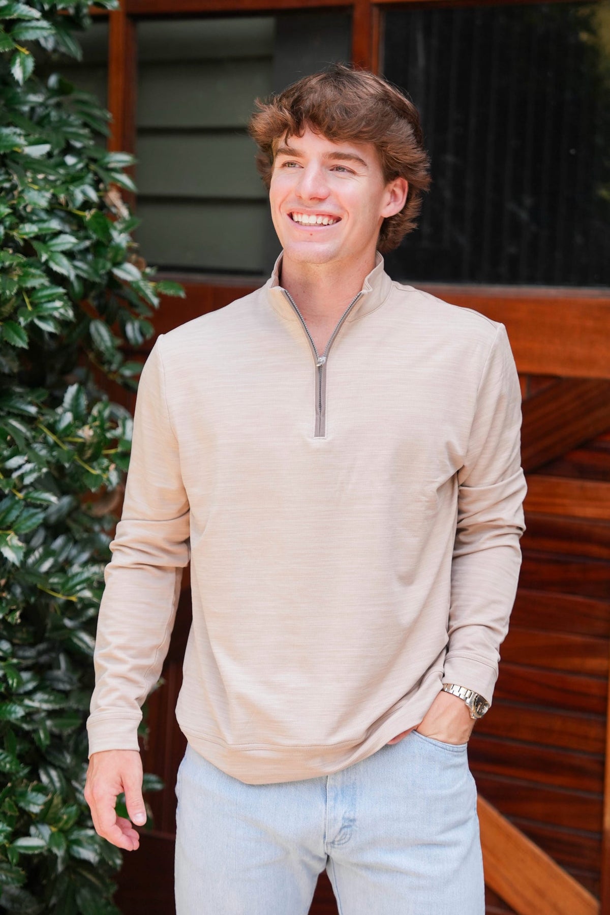 A smiling young man wearing a light beige long-sleeve quarter-zip pullover and light blue jeans, standing outdoors in front of a wooden door and green foliage.
