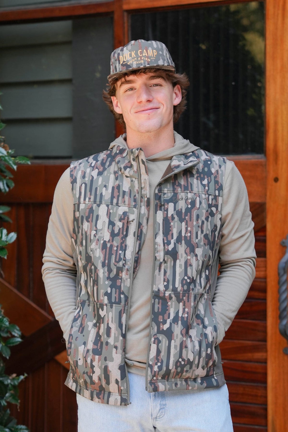 A man wearing a camouflage patterned zip-up vest over a beige hoodie and matching cap, standing in front of a wooden door.