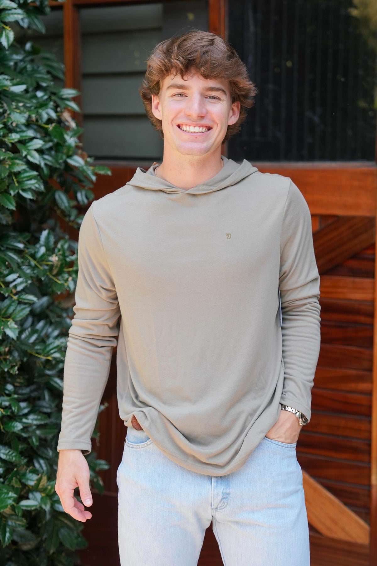 A smiling young man with brown hair wearing a light olive-beige long-sleeve hooded shirt and light blue jeans, standing outdoors in front of a wooden door and green foliage.