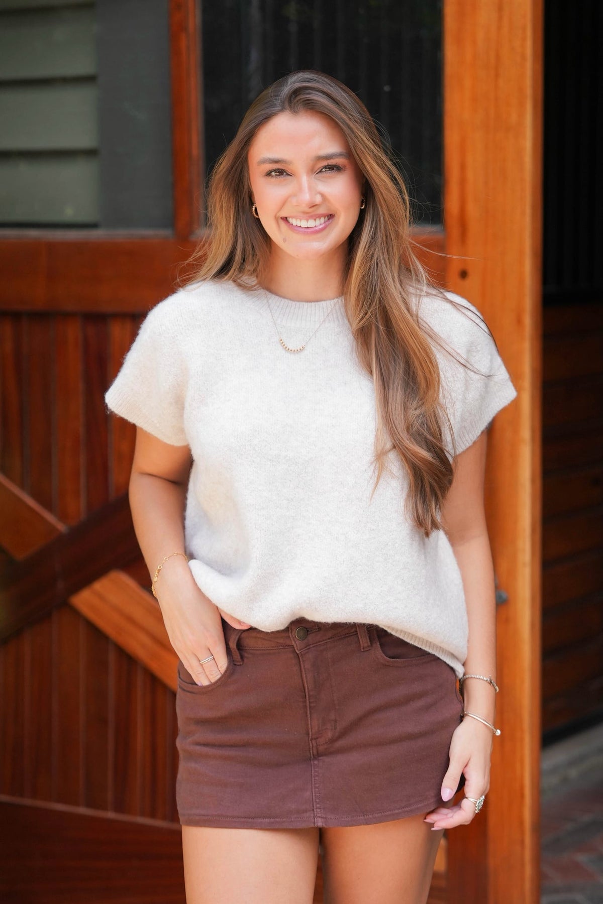A woman smiling and posing in front of a rustic wooden door, wearing a fuzzy off-white short-sleeve t-shirt and a brown mini skirt.