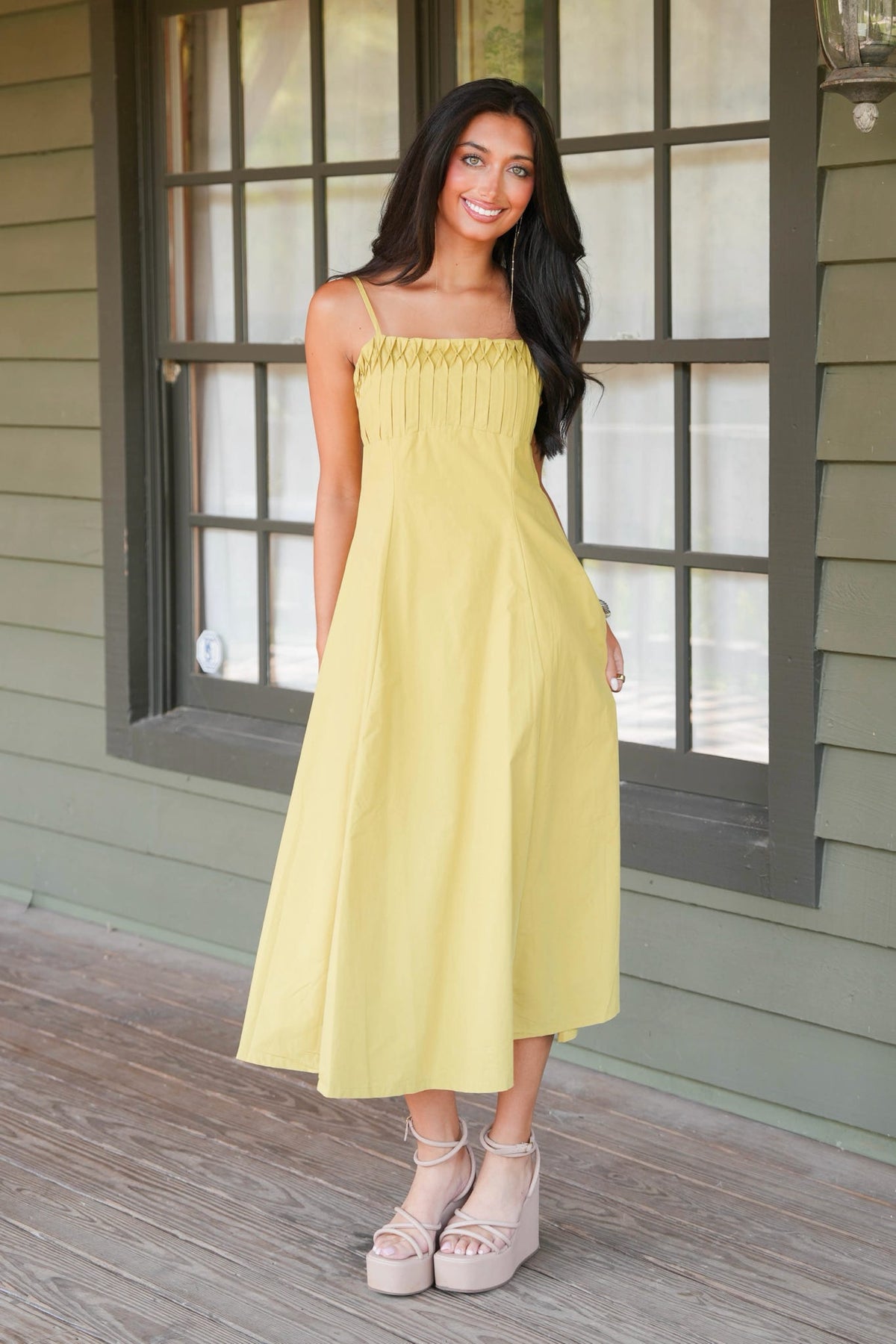 A woman modeling a light yellow midi dress with thin straps and a smocked bodice, standing on a wooden deck in front of a house with green siding and dark-framed windows.