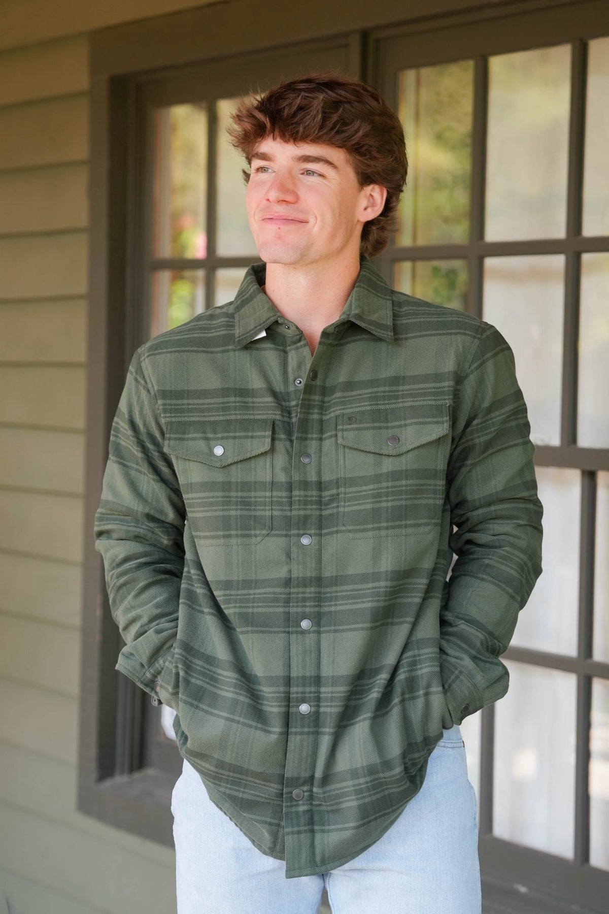 A man wearing an olive green plaid button-down shirt and light blue jeans, standing on a porch in front of a window.