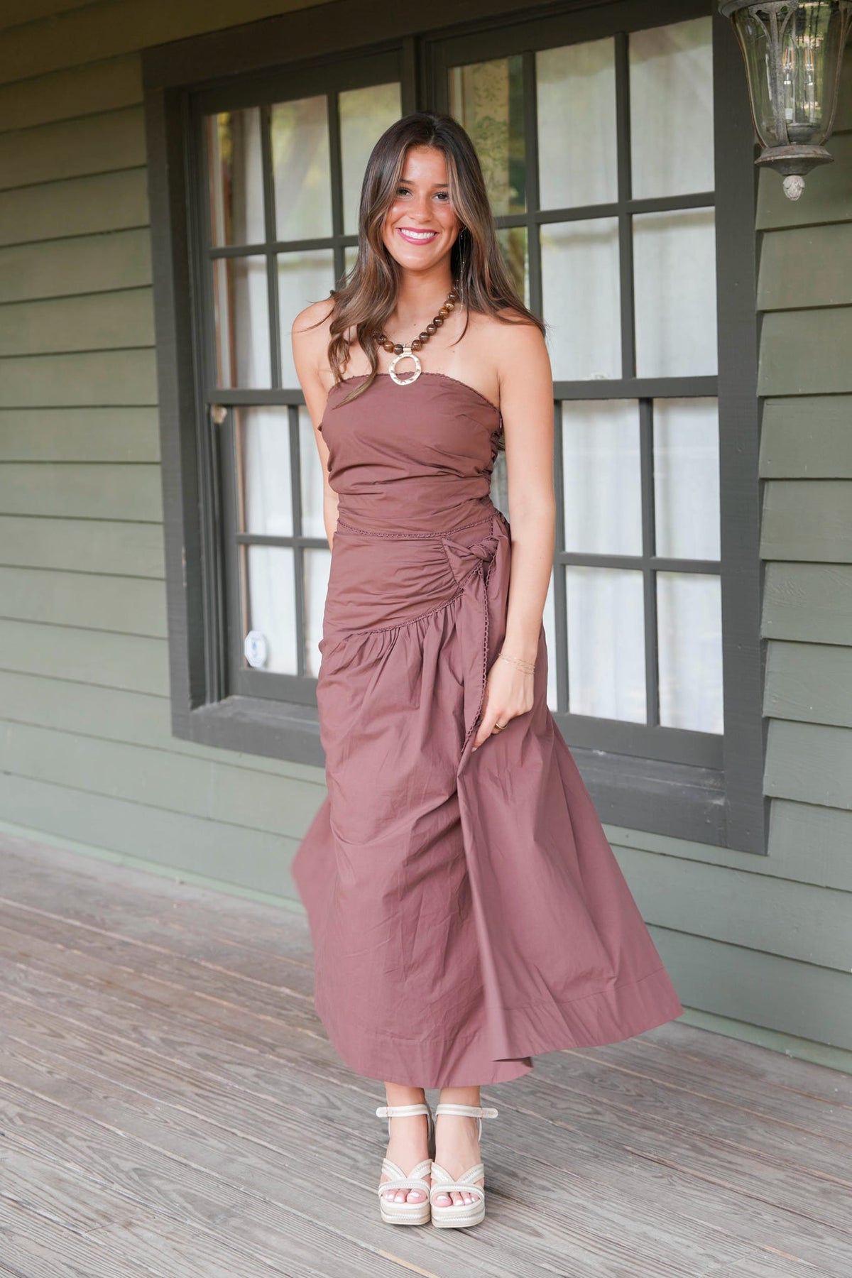 A woman models a strapless dusty rose maxi dress with a gathered ruched bodice and a flowing skirt, standing on a wooden porch in front of a green house with a window.
