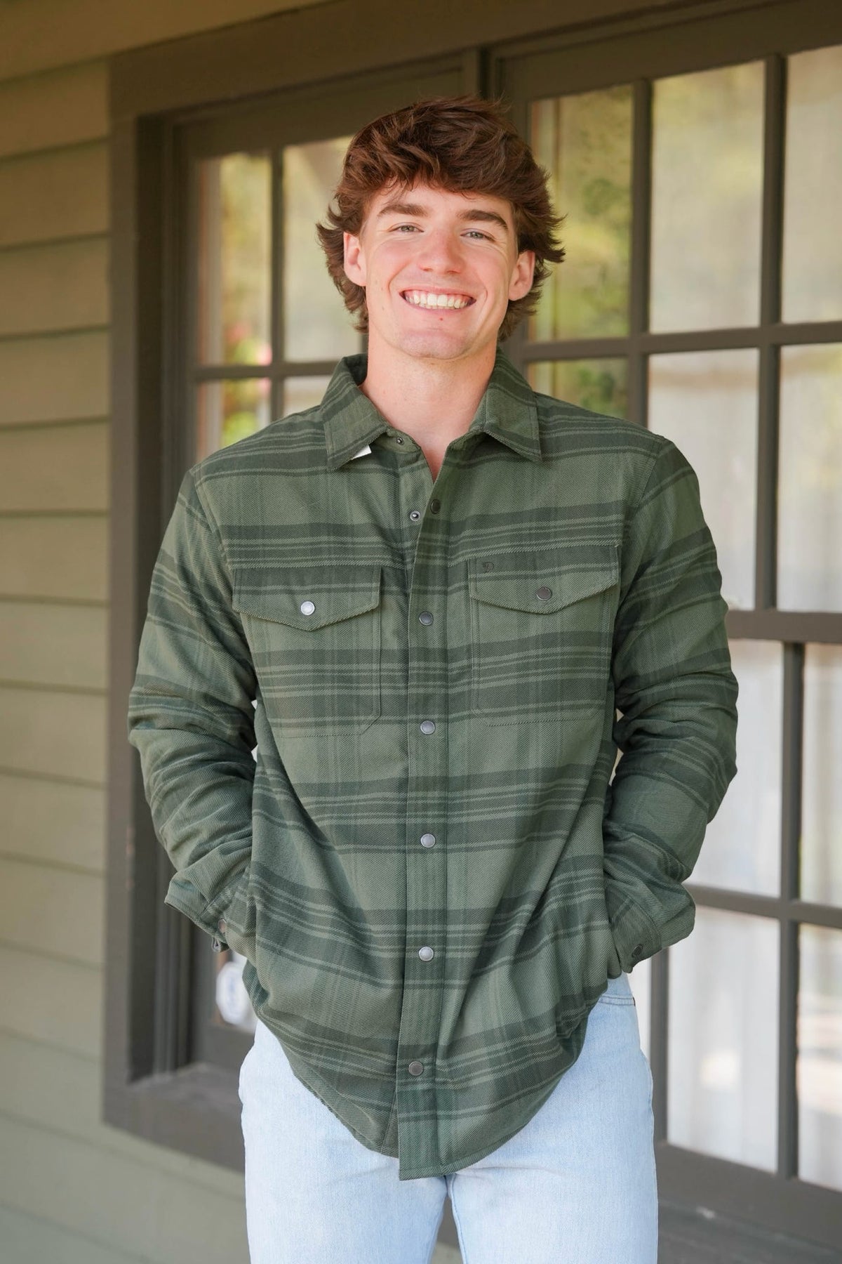 A smiling young man wearing an olive green long-sleeve plaid button-up shirt and light blue jeans, standing in front of a house window.