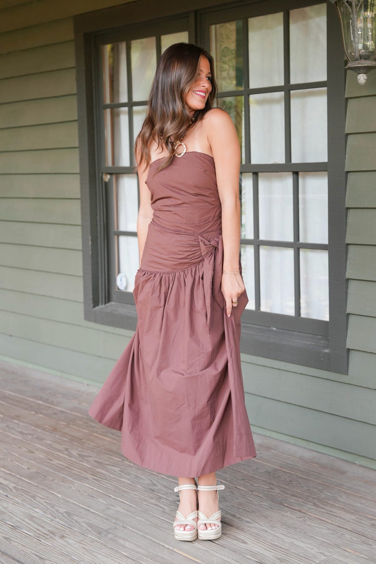 A woman wearing a dusty rose strapless midi dress with a gathered waist and side tie, standing on a wooden porch in front of a green house with a window.