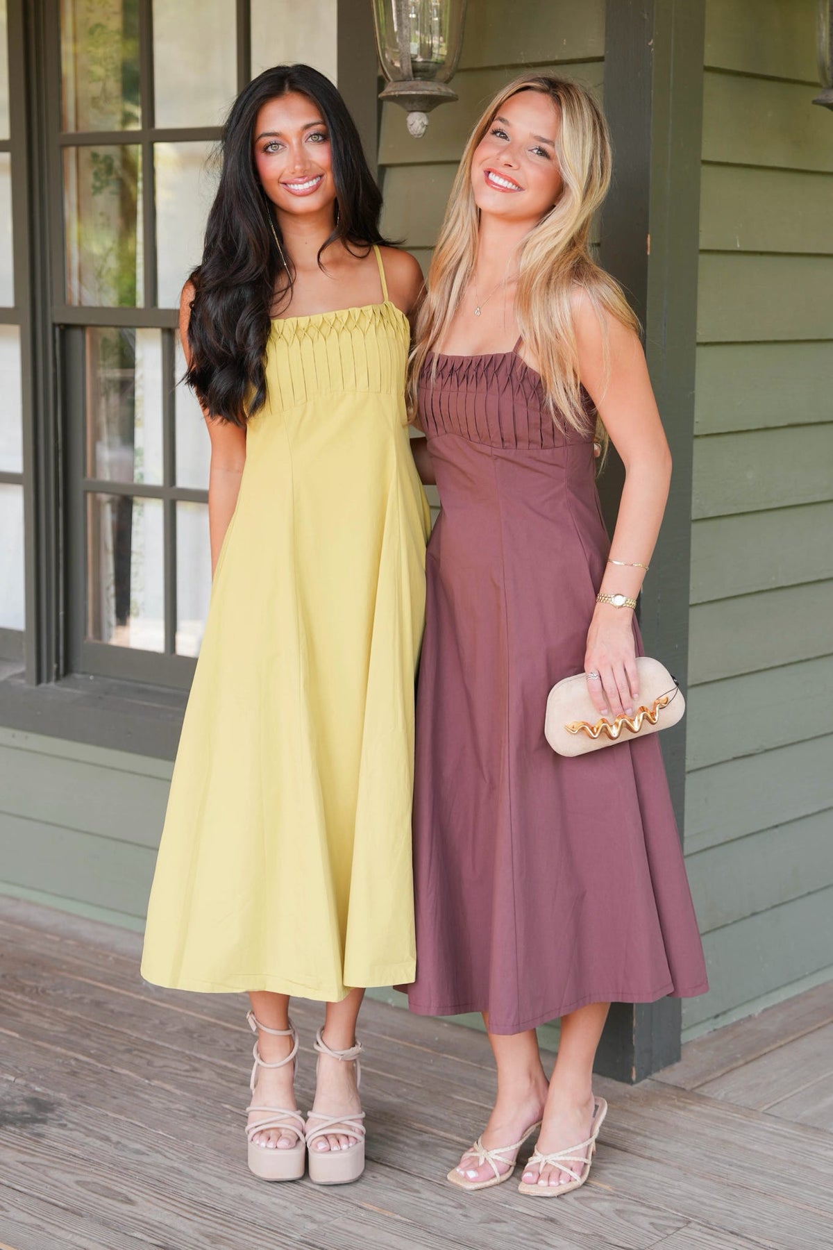 Two women posing on a porch wearing matching midi-length sleeveless dresses in different colors: one is a soft pastel yellow and the other is a dusty mauve-purple. The woman on the right holds a small beige clutch with gold studs.