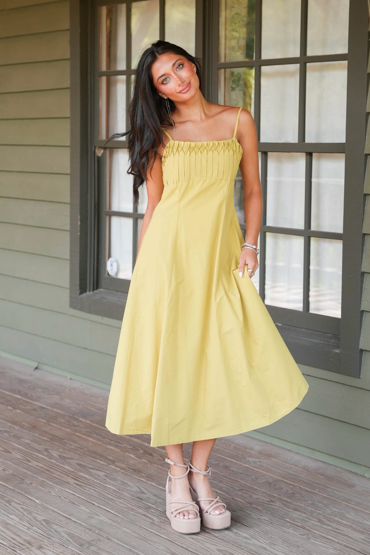 A woman modeling a light yellow midi dress with spaghetti straps and a gathered smocked bodice, standing on a wooden porch in front of a green house with a window.