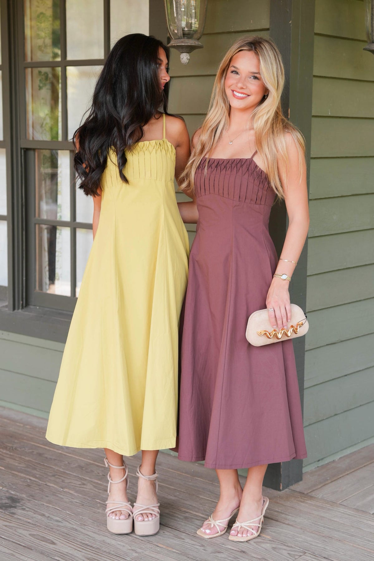 Two women standing on a porch wearing sleeveless midi dresses, one in pale yellow and the other in dusty mauve, with one holding a small beige clutch.