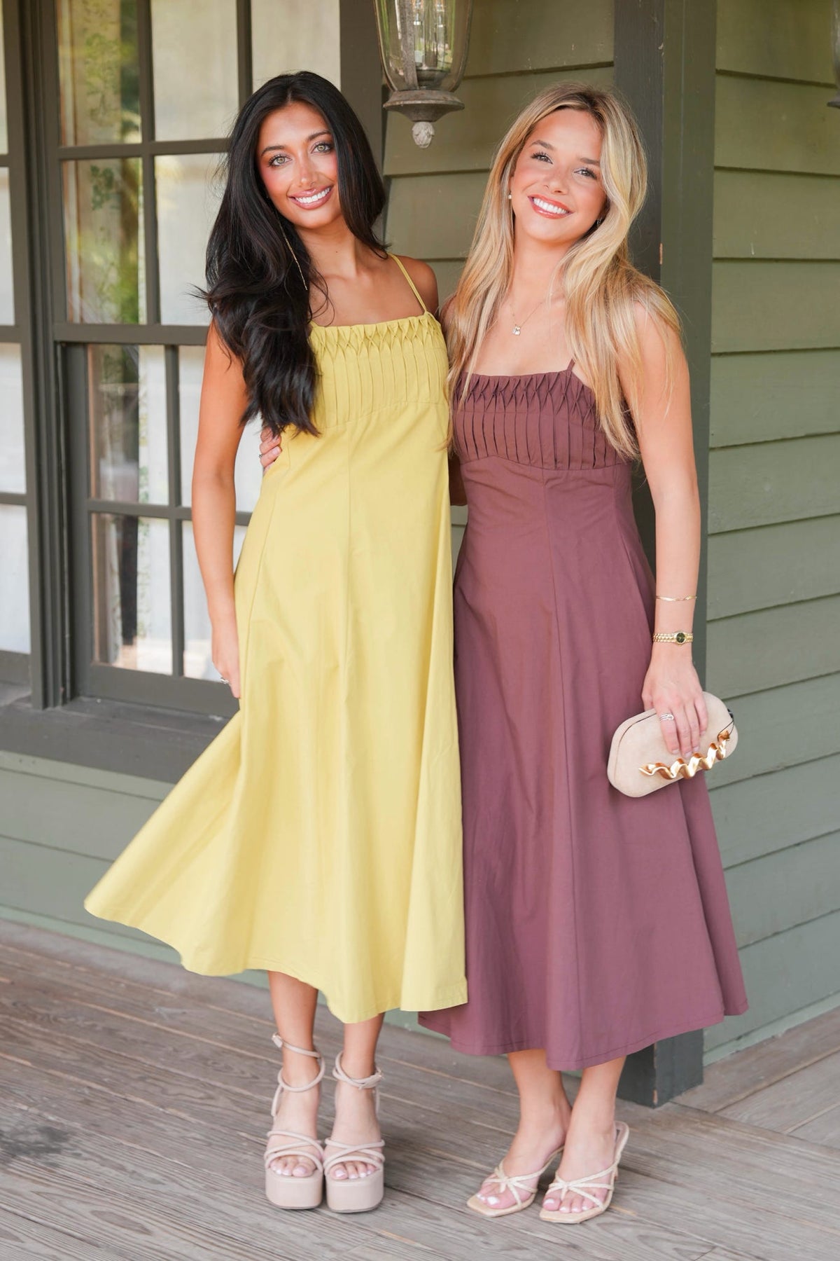 Two women posing on a porch wearing matching midi-length sleeveless dresses in different colors: one is a soft pastel yellow and the other is a dusty mauve-purple. The woman on the right holds a small beige clutch with gold studs.