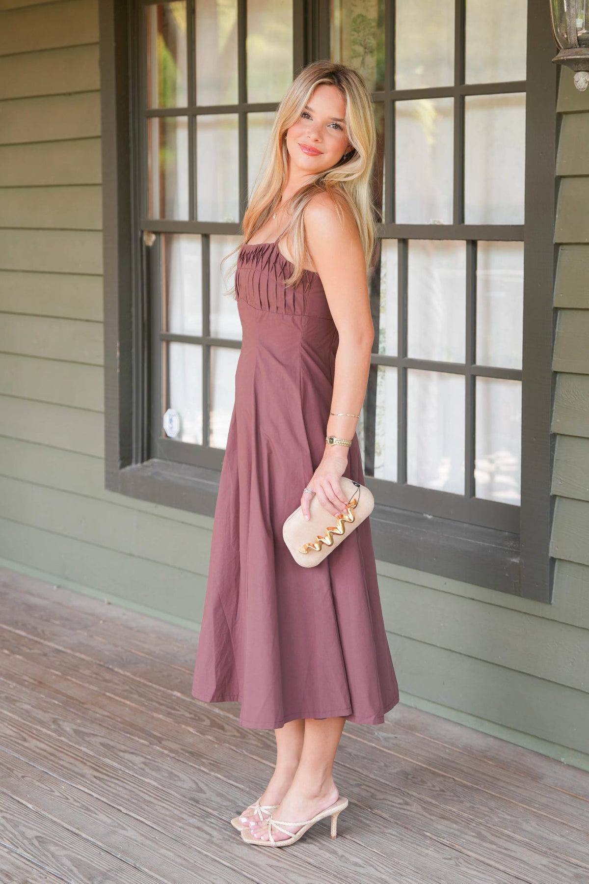 A woman stands on a porch wearing a sleeveless dusty mauve midi dress with a ruched bodice and flowing skirt. She is holding a small cream clutch with gold studs and wearing clear high-heeled mules.