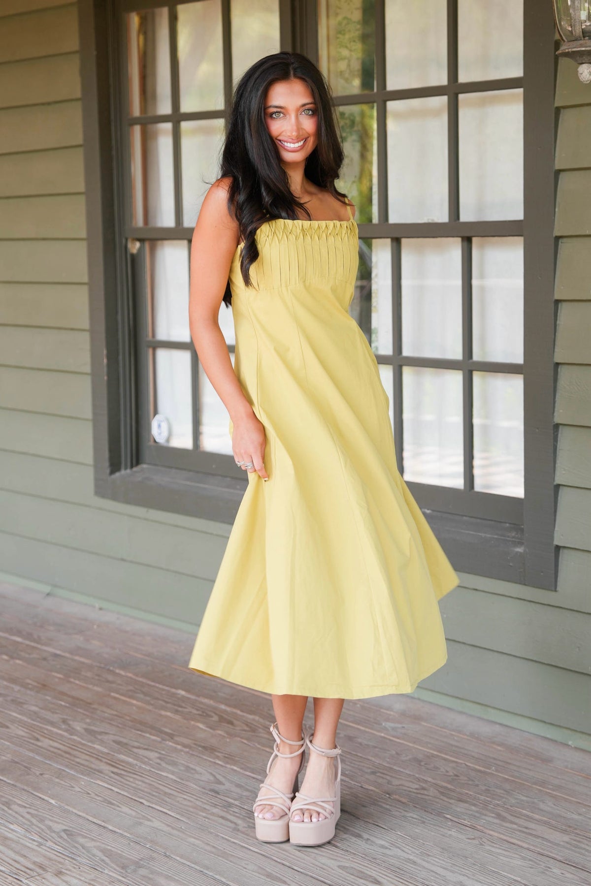 A woman wearing a strapless yellow midi dress and beige platform sandals, standing on a wooden porch in front of a gray-framed window.