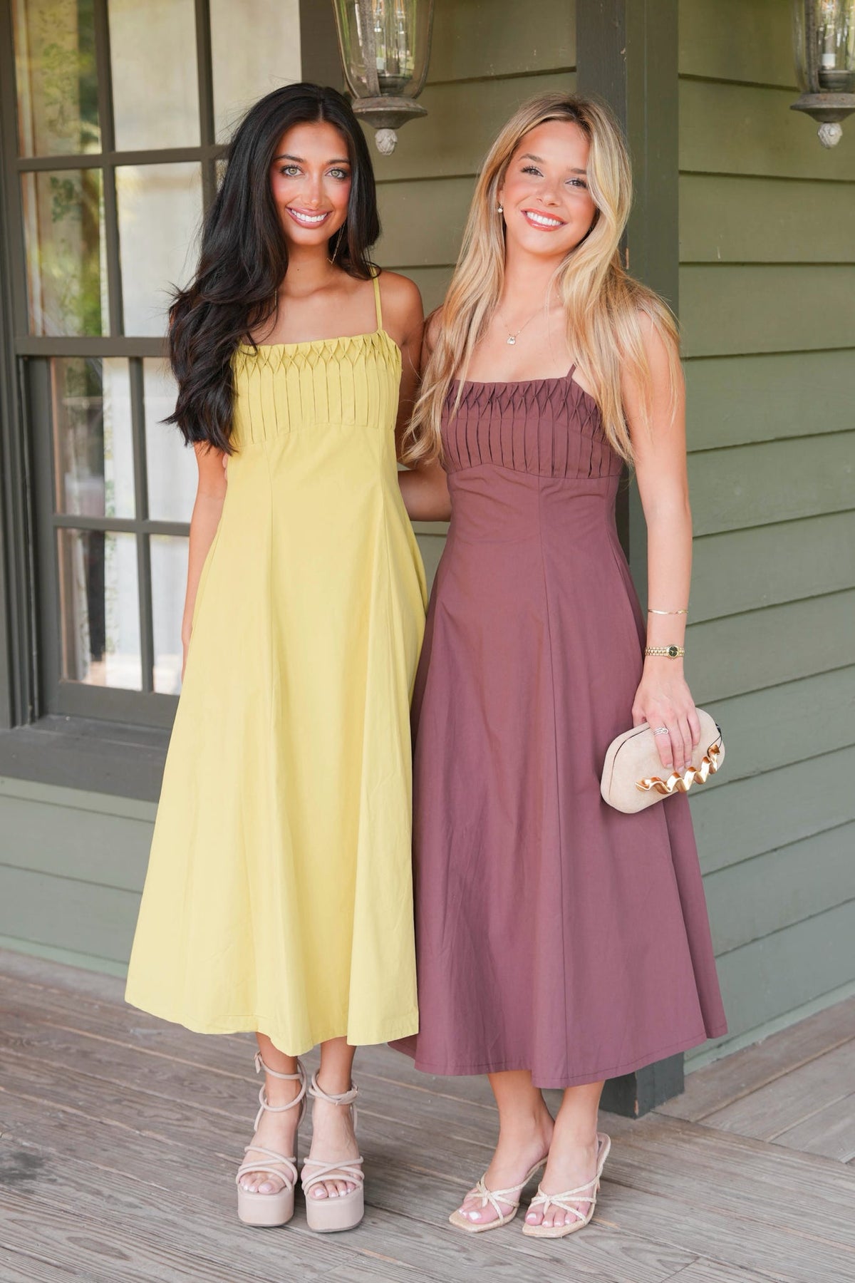 Two women posing on a porch wearing matching midi-length spaghetti-strap dresses, one in pale yellow and the other in dusty mauve, with neutral platform sandals.
