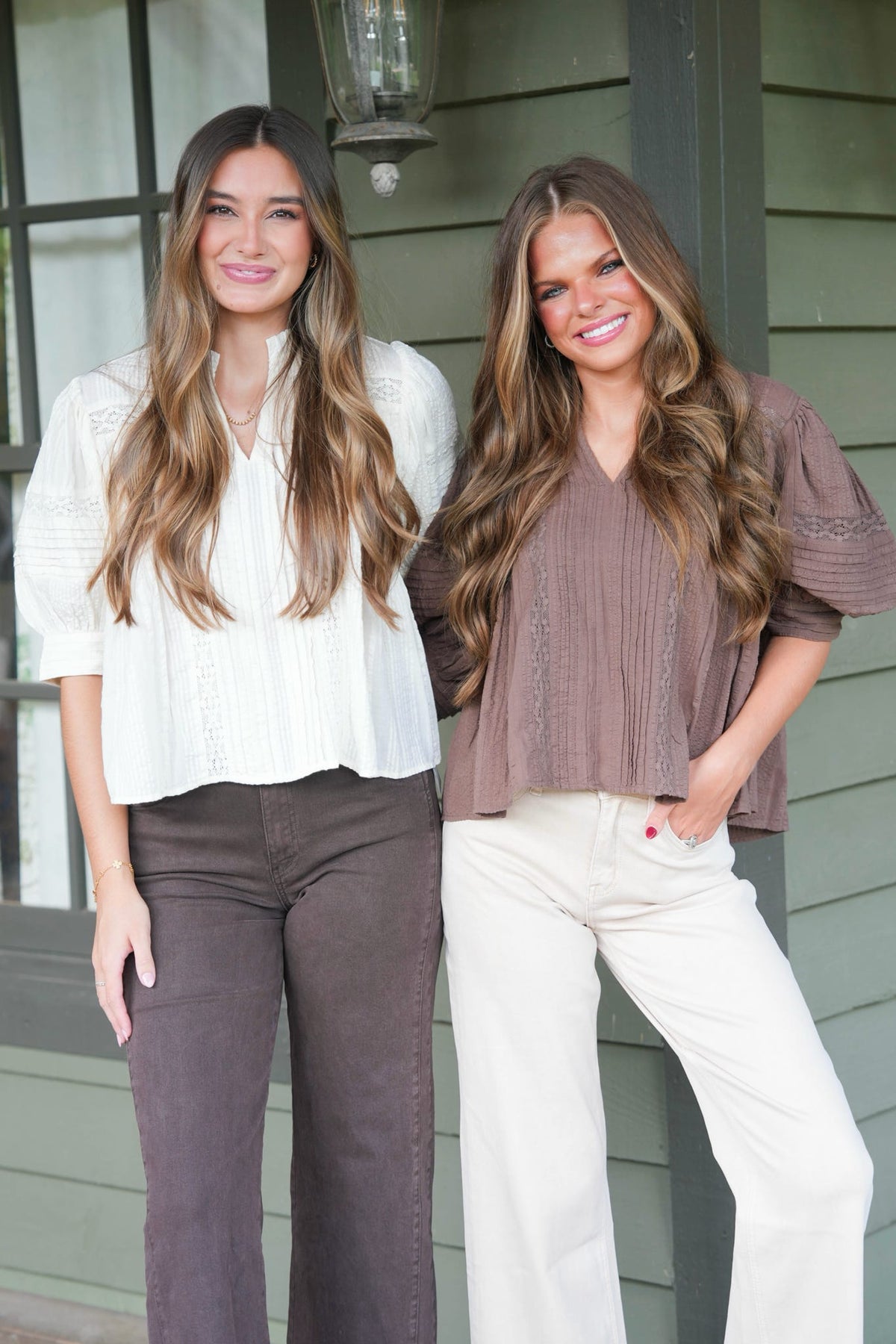Two women standing side-by-side in front of a green house exterior. The woman on the left wears a white textured blouse and dark brown pants.