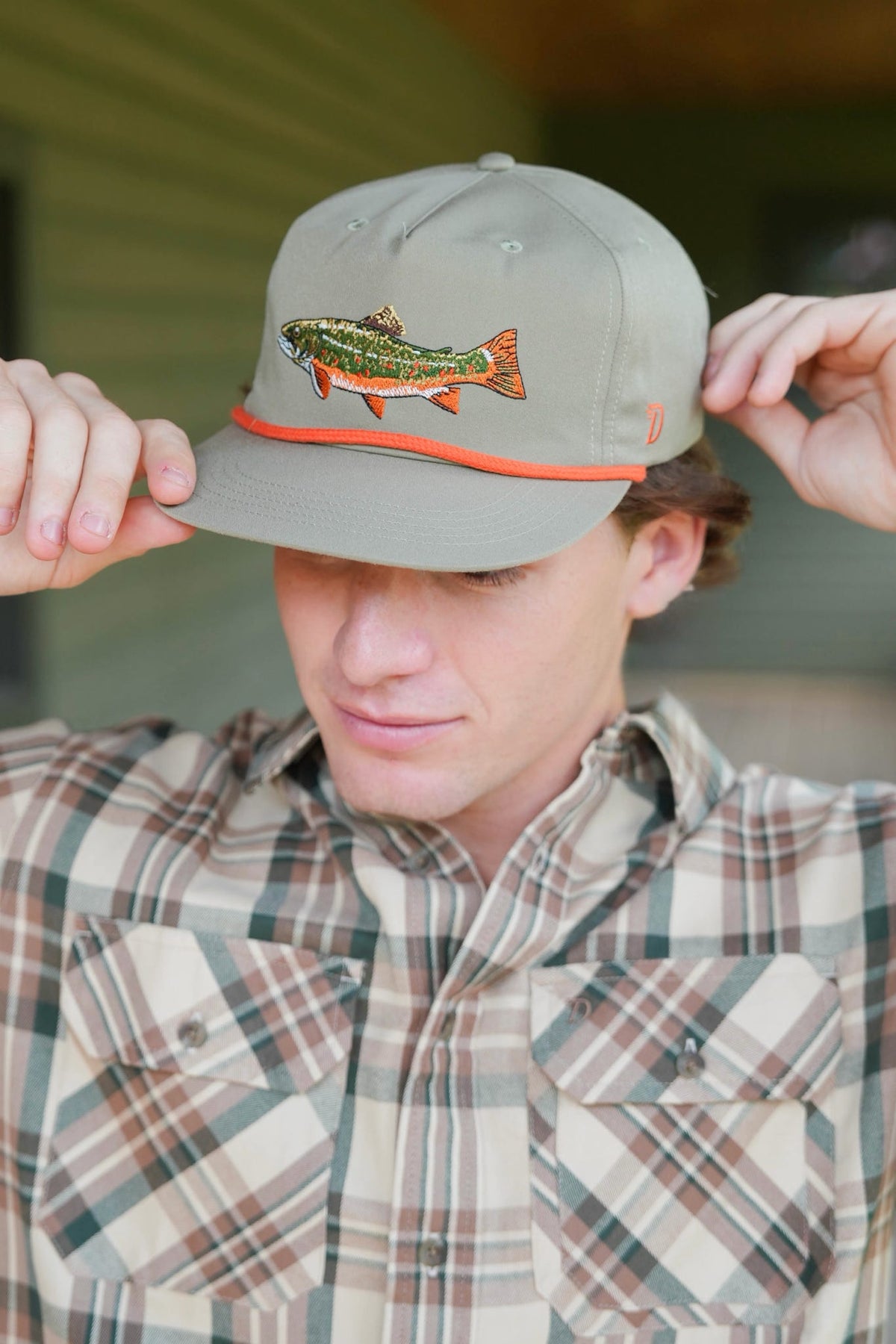 Close-up of a person in a plaid shirt adjusting a sage green cap embroidered with a colorful trout and trimmed with an orange cord.