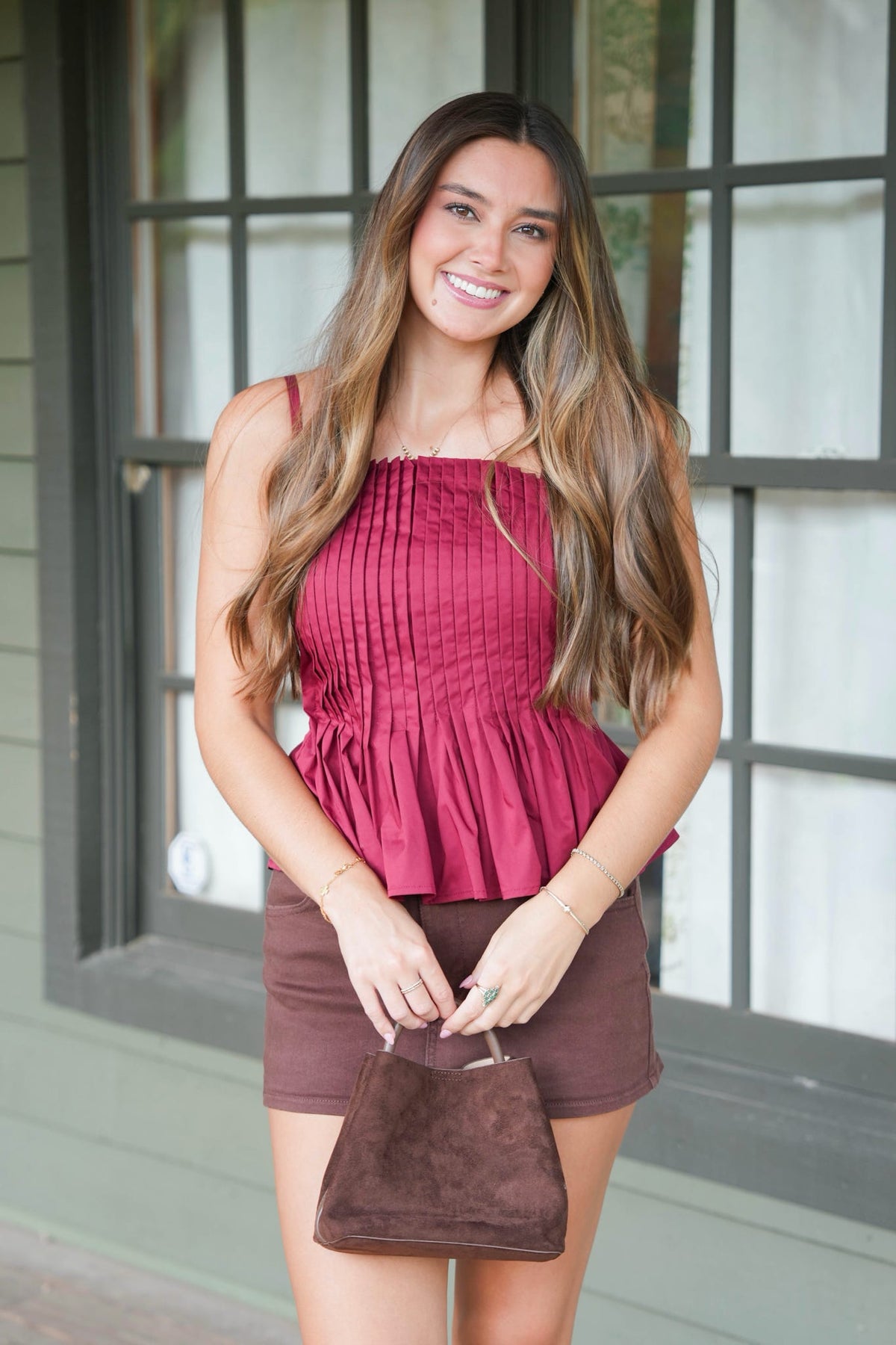 A woman modeling a sleeveless burgundy peplum top and brown shorts, holding a brown suede handbag in front of a windowed exterior wall.