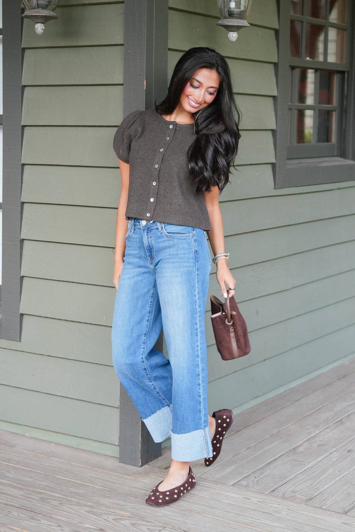 A woman stands on a porch wearing a dark brown short-sleeve cropped top with button details, light blue wide-leg jeans with rolled cuffs, and brown studded flats. She is holding a brown leather handbag.