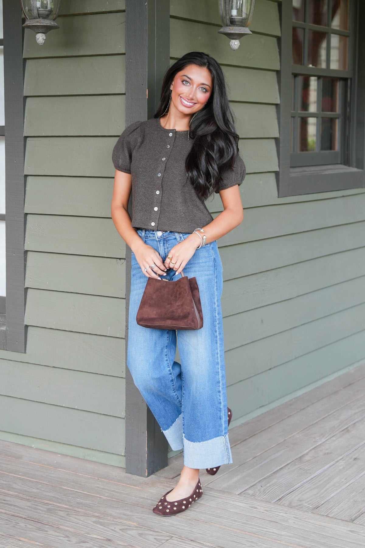 A woman stands on a porch wearing a dark brown short-sleeve cropped top with button details, light blue wide-leg jeans with rolled cuffs, and brown studded flats. She is holding a brown leather handbag.