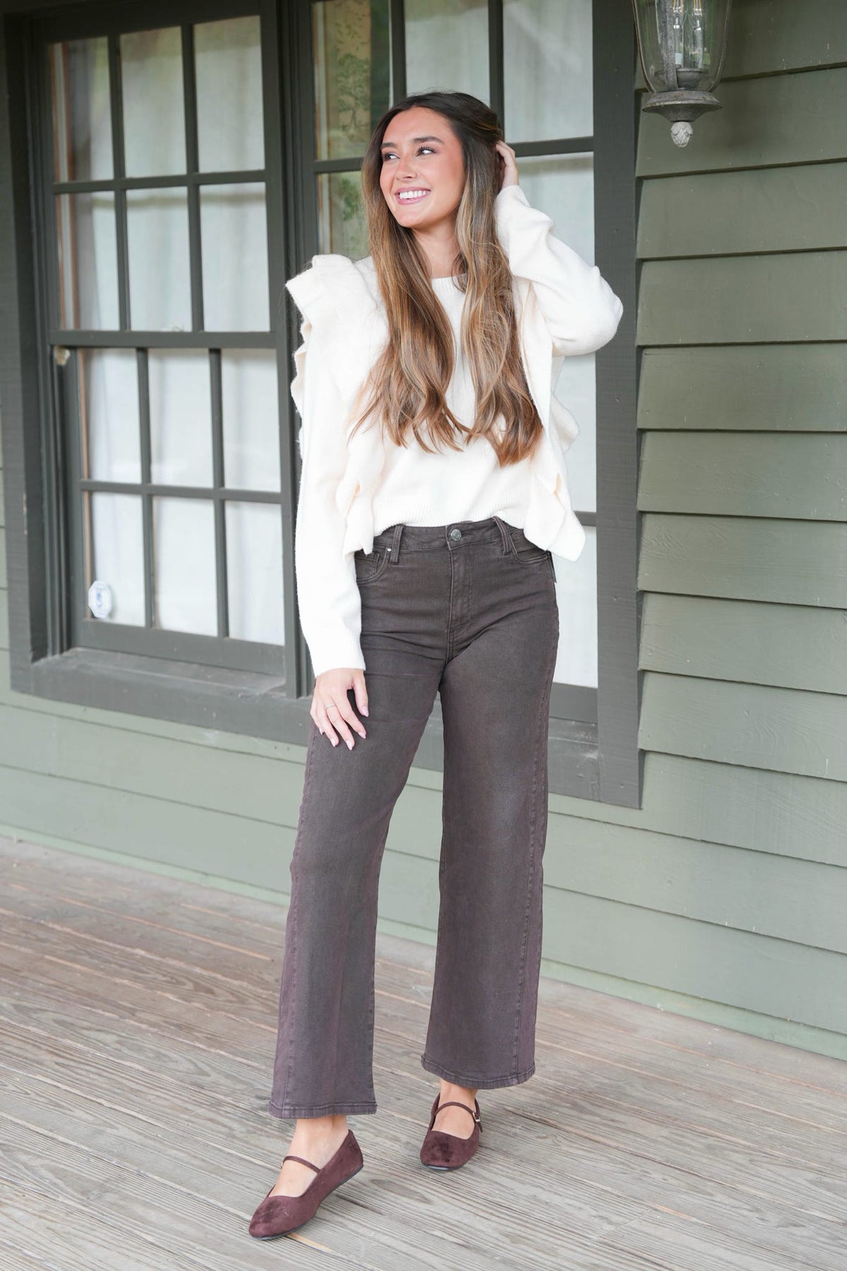 A woman stands on a porch wearing a cream ruffled long-sleeve top, dark brown wide-leg jeans, and burgundy flat shoes.