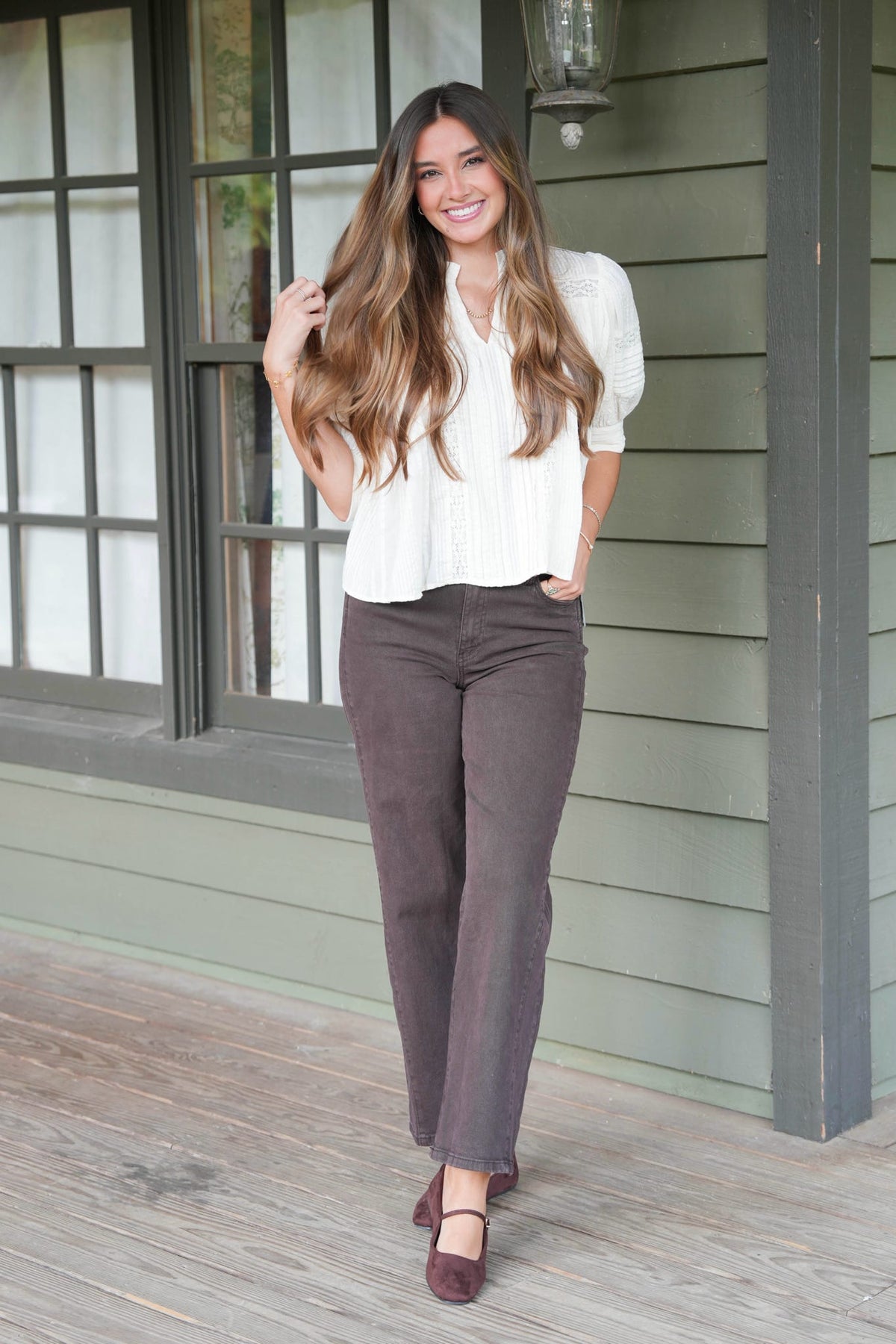A woman stands on a wooden porch wearing a white short-sleeve blouse, dark brown straight-leg pants, and dark brown Mary Jane shoes. She is smiling and posing in front of a house with green siding and dark-framed windows.