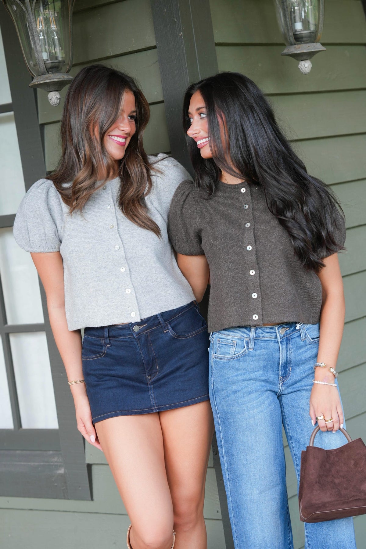 Two women smiling and posing together on a porch. The woman on the left wears a light gray puff-sleeve crop top with a dark denim mini skirt, and the woman on the right wears a dark brown puff-sleeve button-front crop top with light-wash blue jeans.