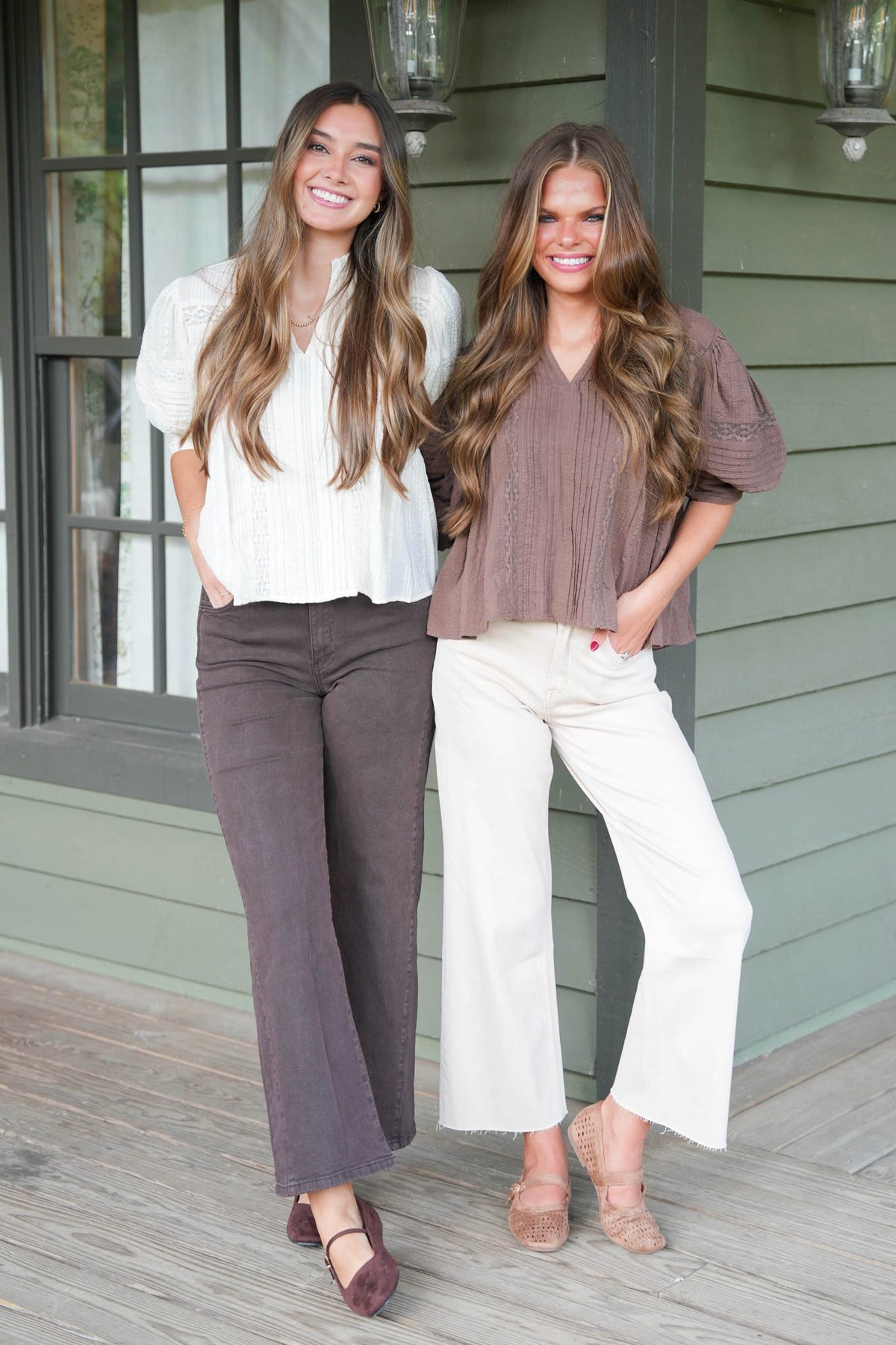 Two women standing on a porch wearing matching blouse-and-pants outfits. The woman on the left wears a white short-sleeve blouse with dark brown wide-leg pants, and the woman on the right wears a taupe short-sleeve blouse with off-white wide-leg pants.
