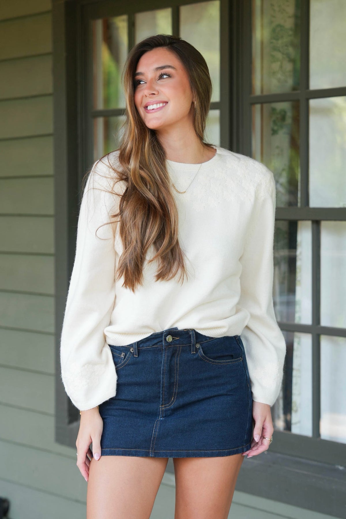 A woman wearing a cream-colored long-sleeve knit sweater and a dark blue denim mini skirt, standing in front of a house window.