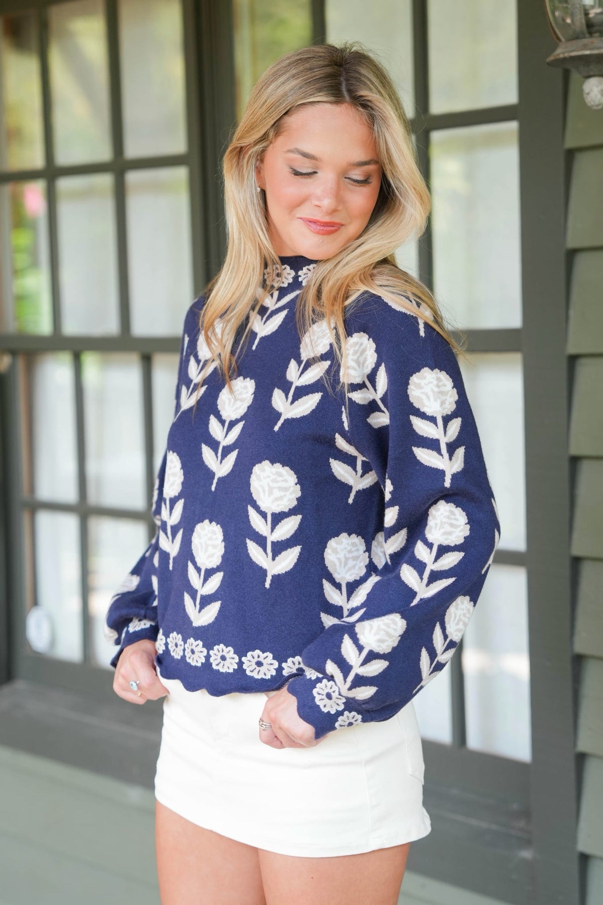 A woman wearing a navy blue long-sleeve knit sweater with a white floral block-print pattern, paired with a white mini skirt, standing in front of a dark-framed window.
