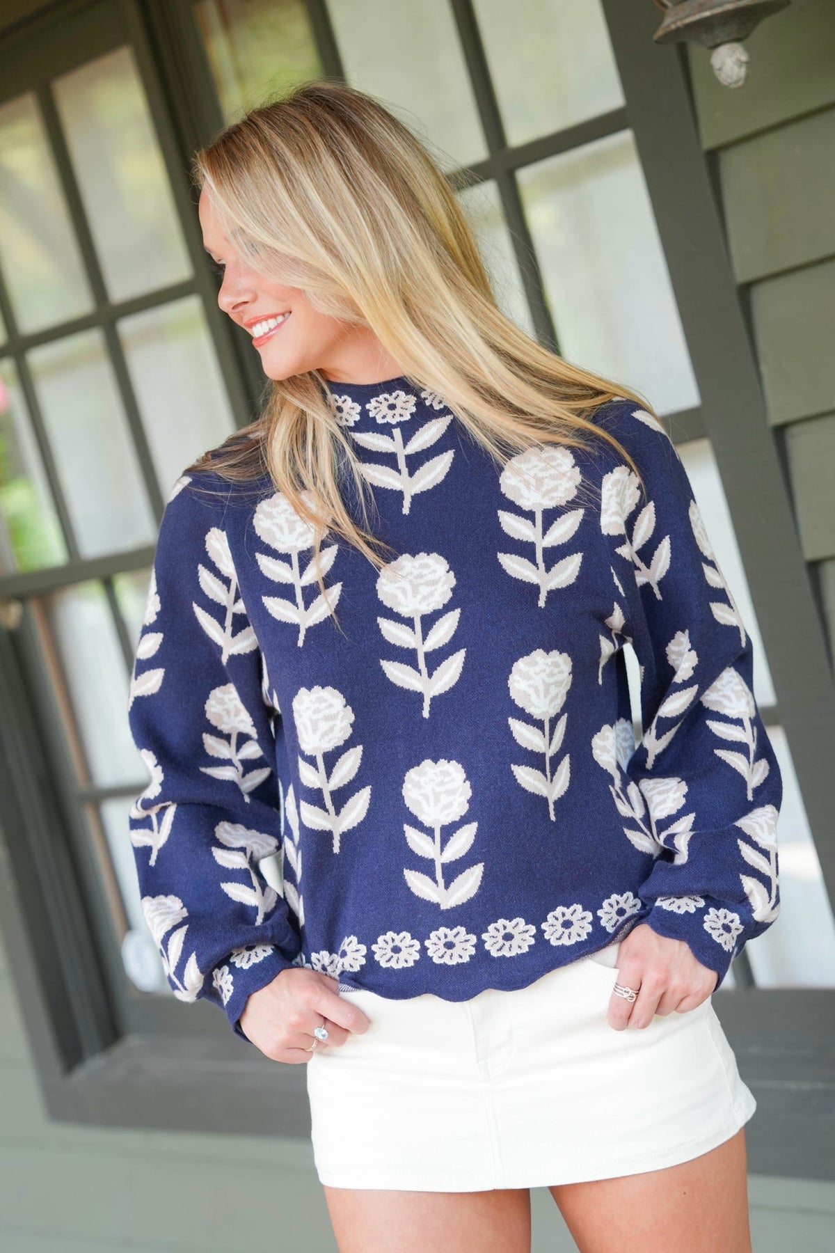 A woman wearing a navy blue knit sweater with a white floral pattern, paired with a white mini skirt, standing in front of a gray paneled window.