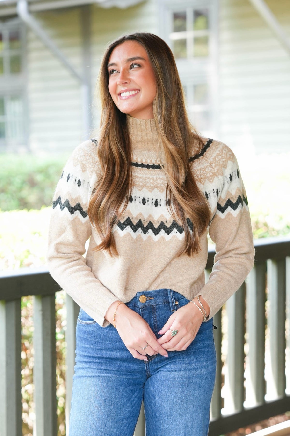 A woman with long brown hair smiling and looking to the side, wearing a beige knit sweater with black and white Nordic-style patterns and blue jeans, standing on a porch.