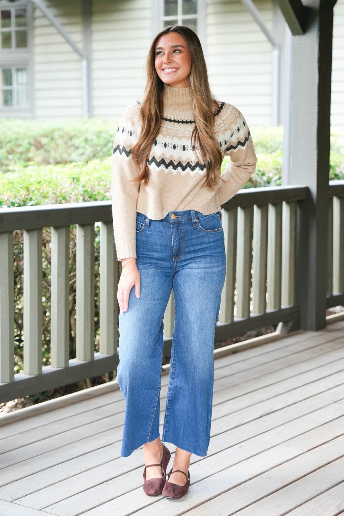 A woman stands on a wooden porch wearing a beige knit turtleneck sweater with a dark geometric pattern across the chest and shoulders, paired with blue wide-leg cropped jeans and brown flats.