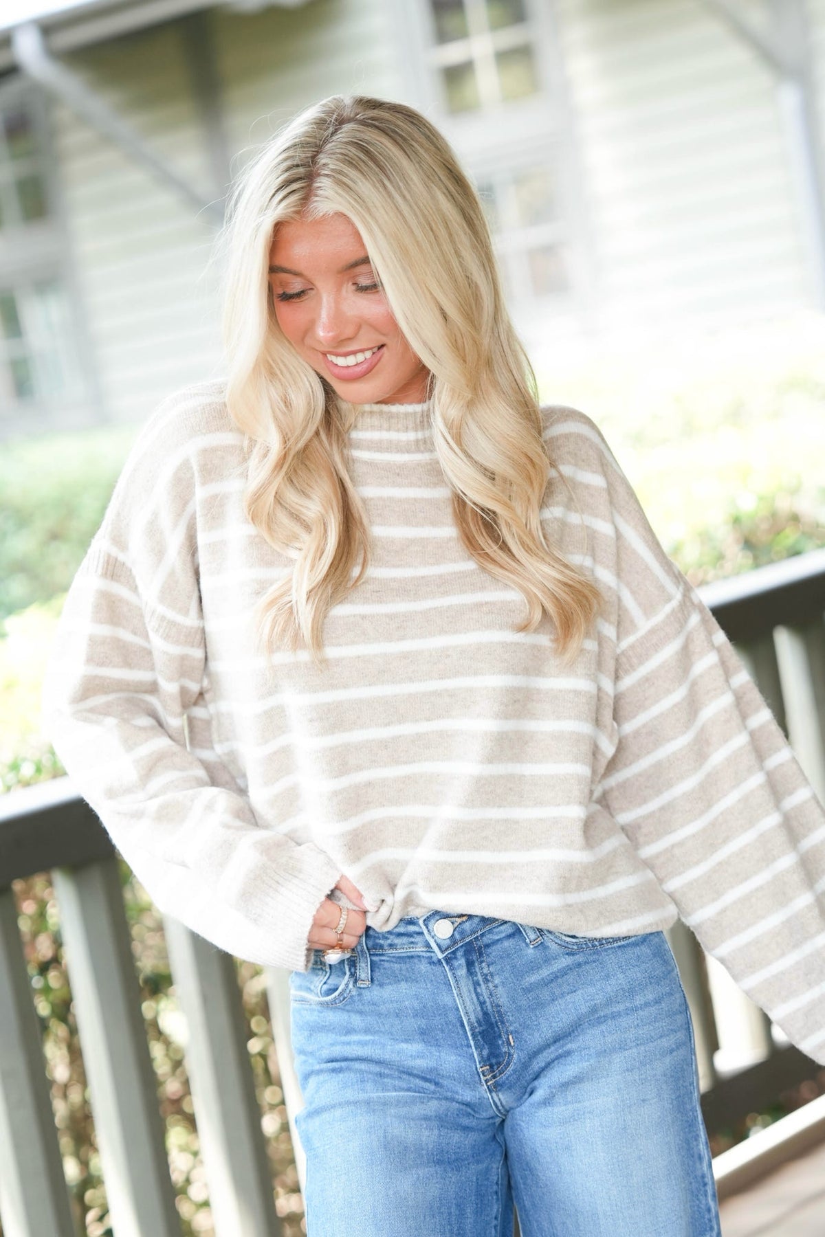 A woman with long blonde hair wearing a cream and beige horizontally striped knit sweater and blue jeans, standing on a porch.