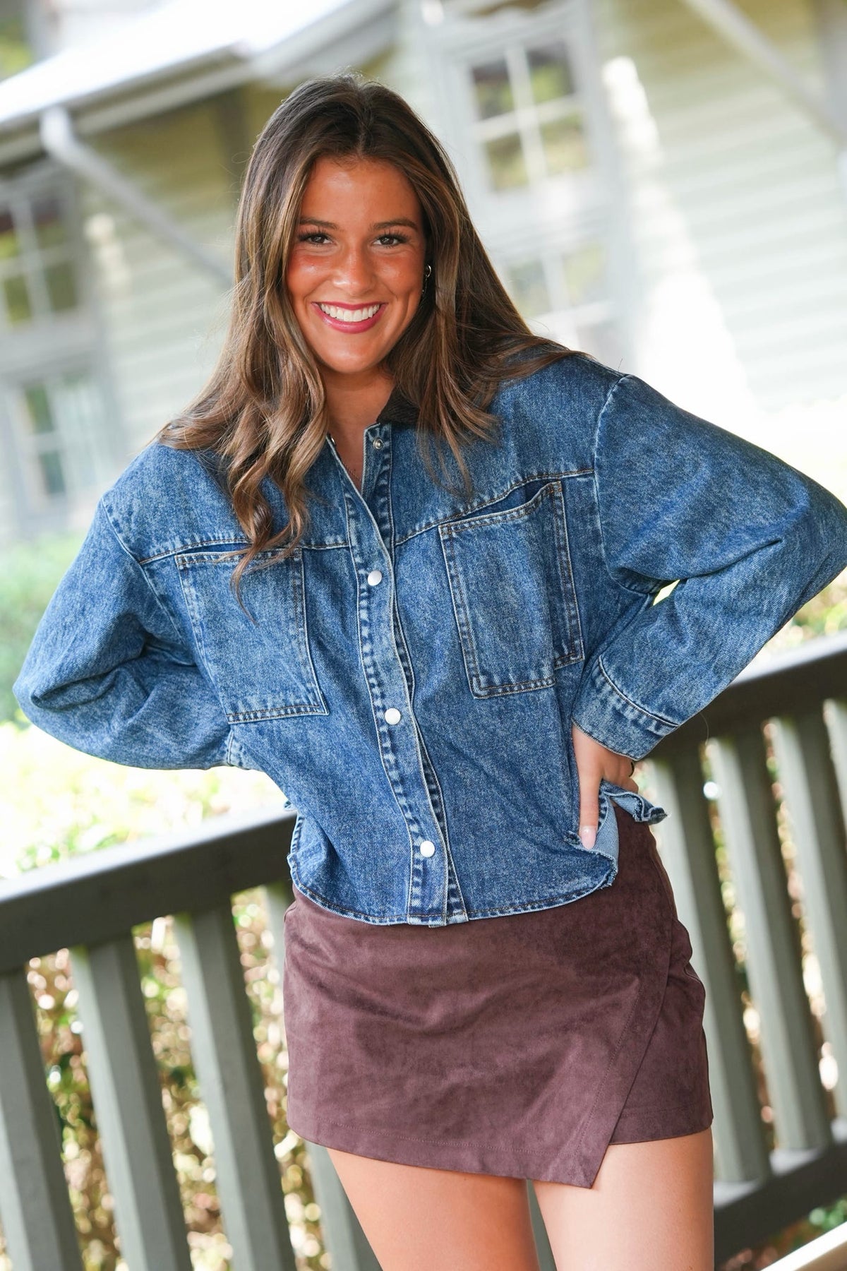A woman smiling on a porch wearing a blue denim button-up shirt and a brown mini skirt.