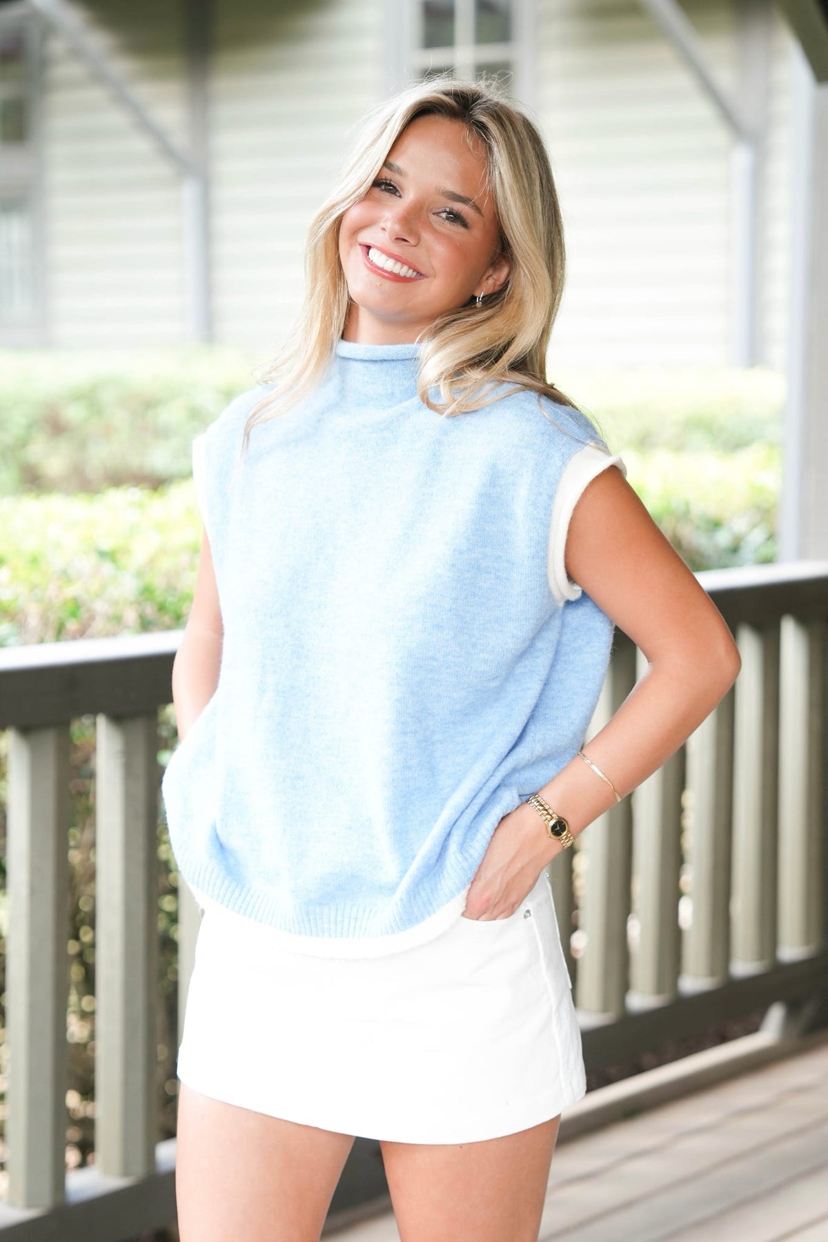 A smiling blonde woman stands on a porch wearing a light blue sleeveless mock-neck knit sweater vest over a white top and a white mini skirt.