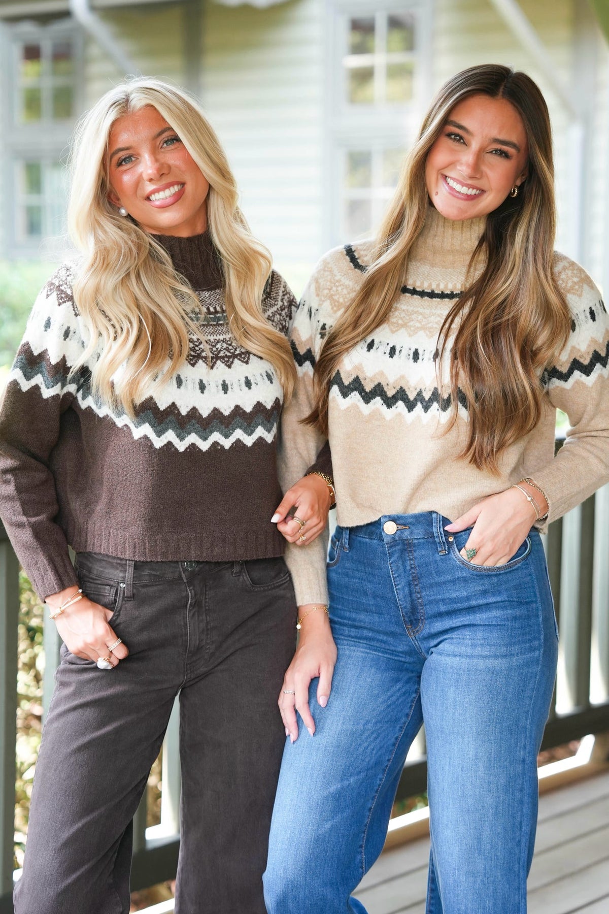 Two women standing on a porch wearing matching knit turtleneck sweaters with a Nordic-style pattern. The woman on the left wears a dark brown and cream sweater with dark gray pants, and the woman on the right wears a beige and cream sweater with blue jeans.