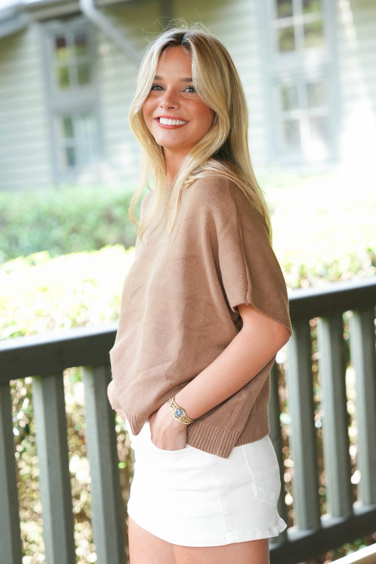 A smiling blonde woman wearing a loose taupe knit short-sleeve top and a white skirt, standing outdoors on a porch with a house and railing in the background.