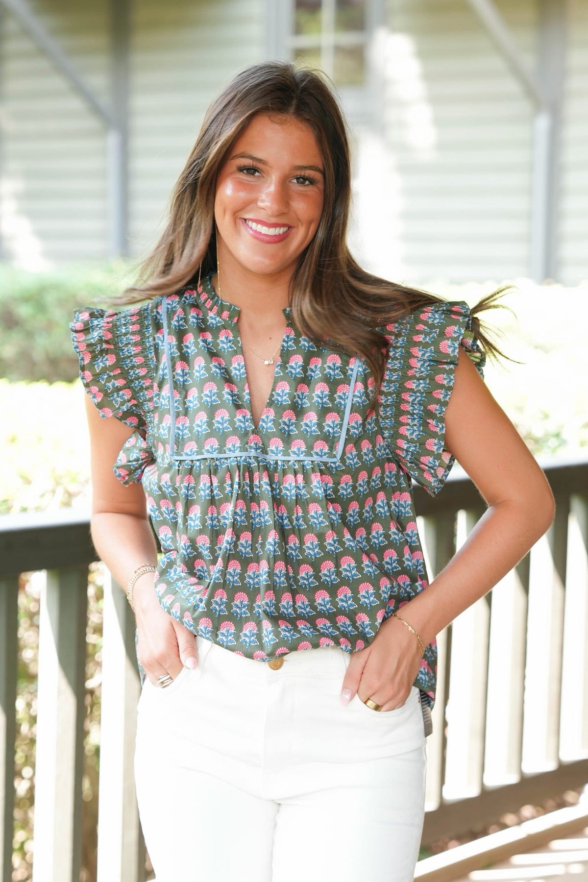 A woman smiling on a porch wearing a short-sleeve ruffled blouse with a colorful floral pattern in teal, pink, and orange, paired with white pants.